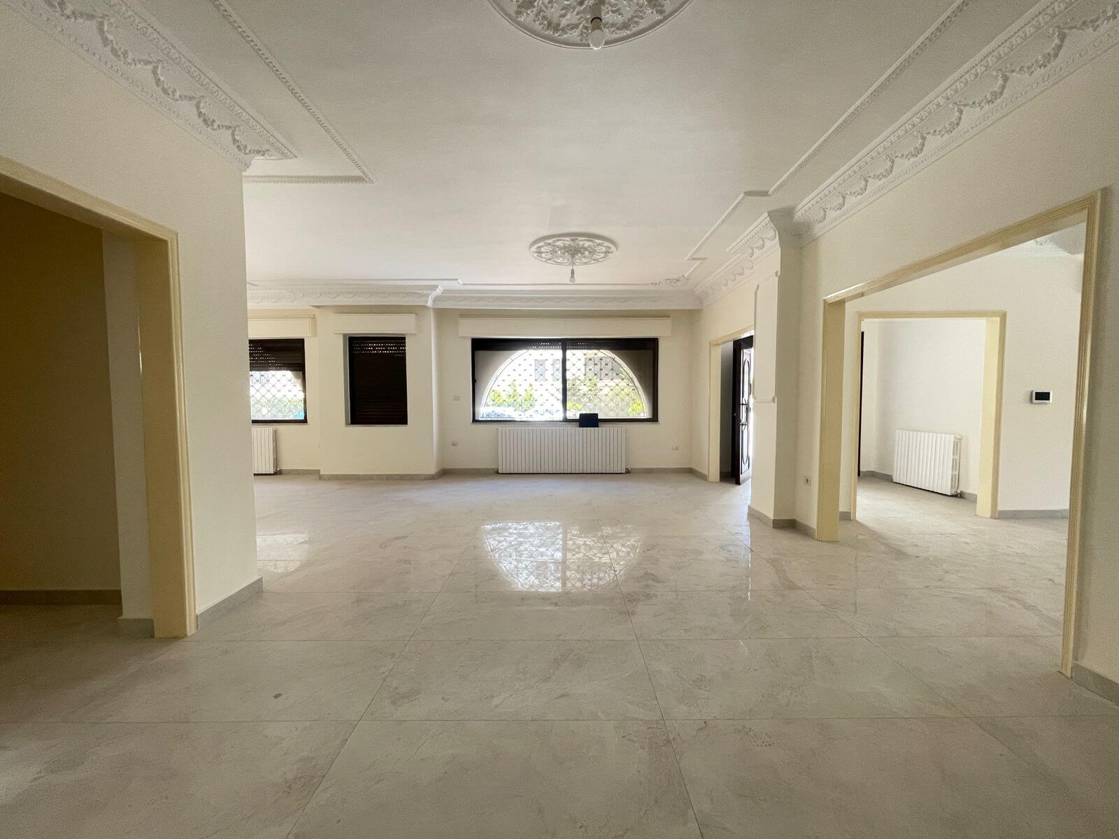 9f89bce1-0ccc-4fd5-9916-615476b452b2 Bright, empty living area with marble floor, ornate ceiling molding, and a large arched window with a metal grille; radiator beneath the window and doorways on the sides