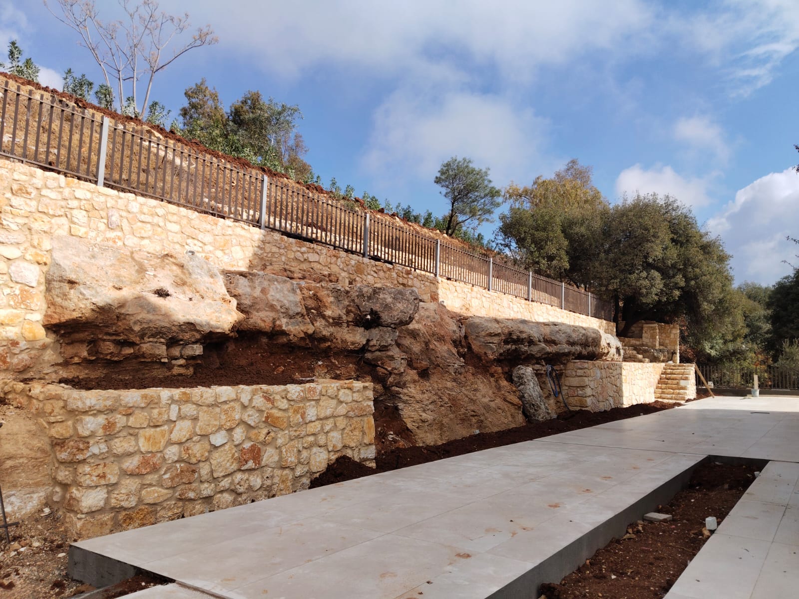 WhatsApp Image 2026-04-08 at 23510 PM 10 Stone retaining wall and railing along a hillside with trees, above a wide concrete walkway and steps on the right, under a blue sky.