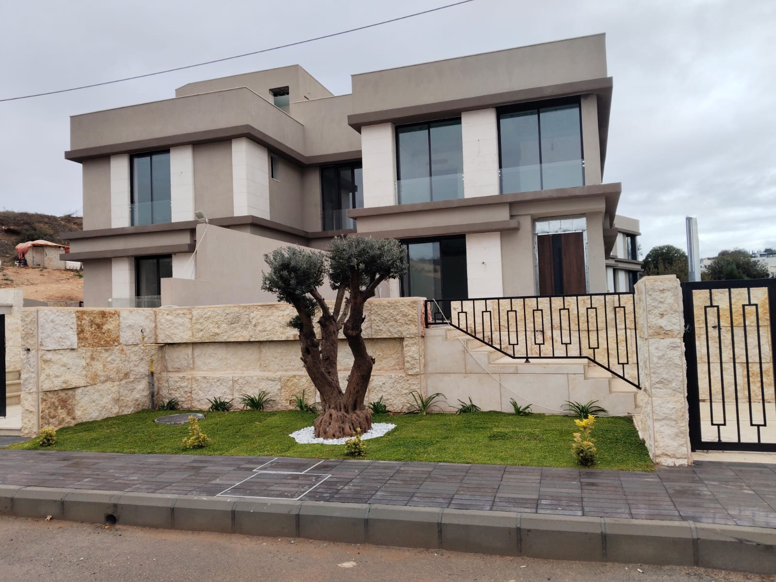 WhatsApp Image 2026-04-08 at 23510 PM 11 Modern two-story house with beige stucco, large glass windows, and a stone-and-tile fence with a black metal gate.