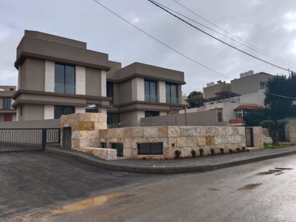 Modern beige house with large windows and stone wall along a curved driveway, gated entry and overhead power lines under a cloudy sky.