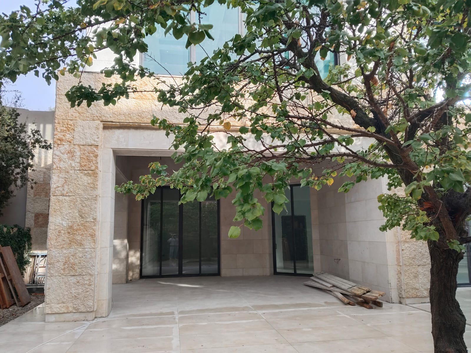 WhatsApp Image 2026-04-08 at 23510 PM 16 Modern stone building facade with large glass doors, partially shaded by a tree in the foreground.