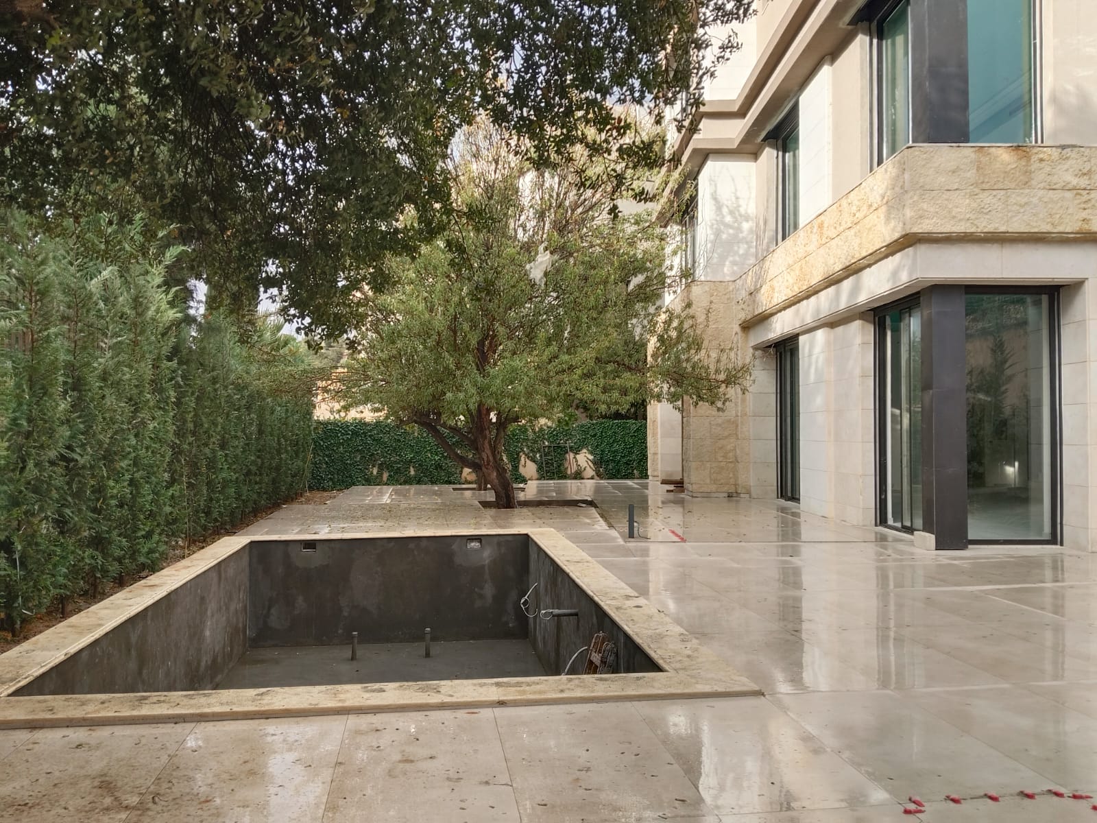 WhatsApp Image 2026-04-08 at 23510 PM 17 Modern beige stone building with large glass doors beside an empty rectangular pool and a tree in a paved courtyard.
