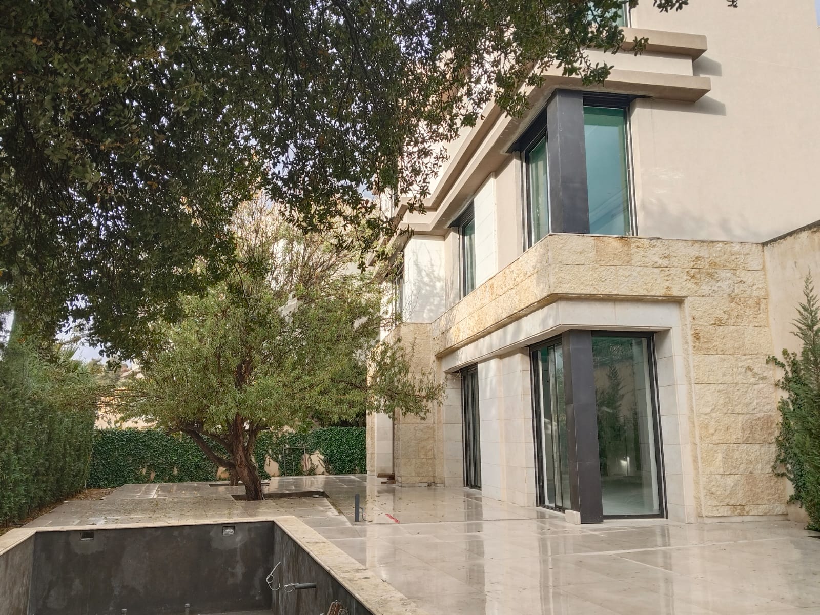 WhatsApp Image 2026-04-08 at 23510 PM 18 Modern beige stone house with large glass doors, surrounded by trees in a landscaped yard and an empty rectangular pool in the foreground.