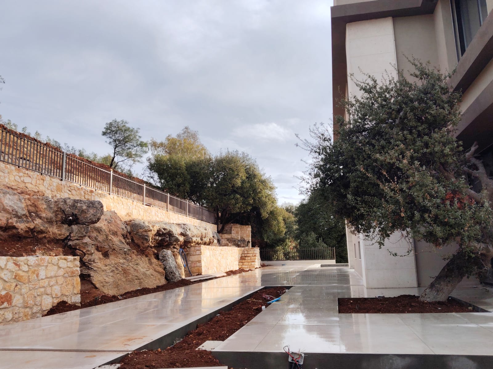 WhatsApp Image 2026-04-08 at 23510 PM 2 Modern building on the right with a tree-filled stone terrace and a wet tiled walkway along a stone wall.