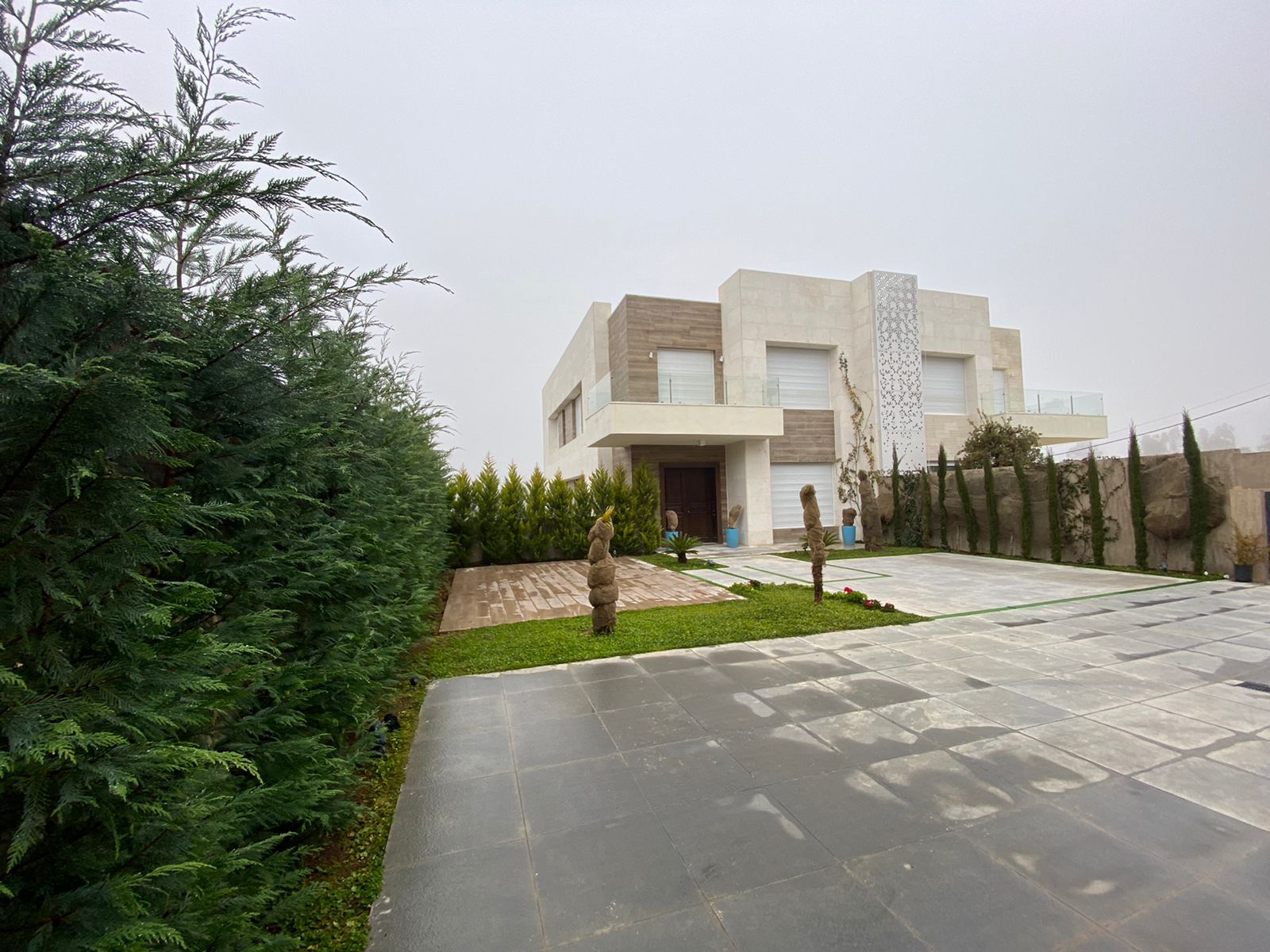 WhatsApp Image 2026-04-08 at 23510 PM 20 Modern two-story house with a stone facade and a tiled driveway, bordered by a dense evergreen hedge on the left.