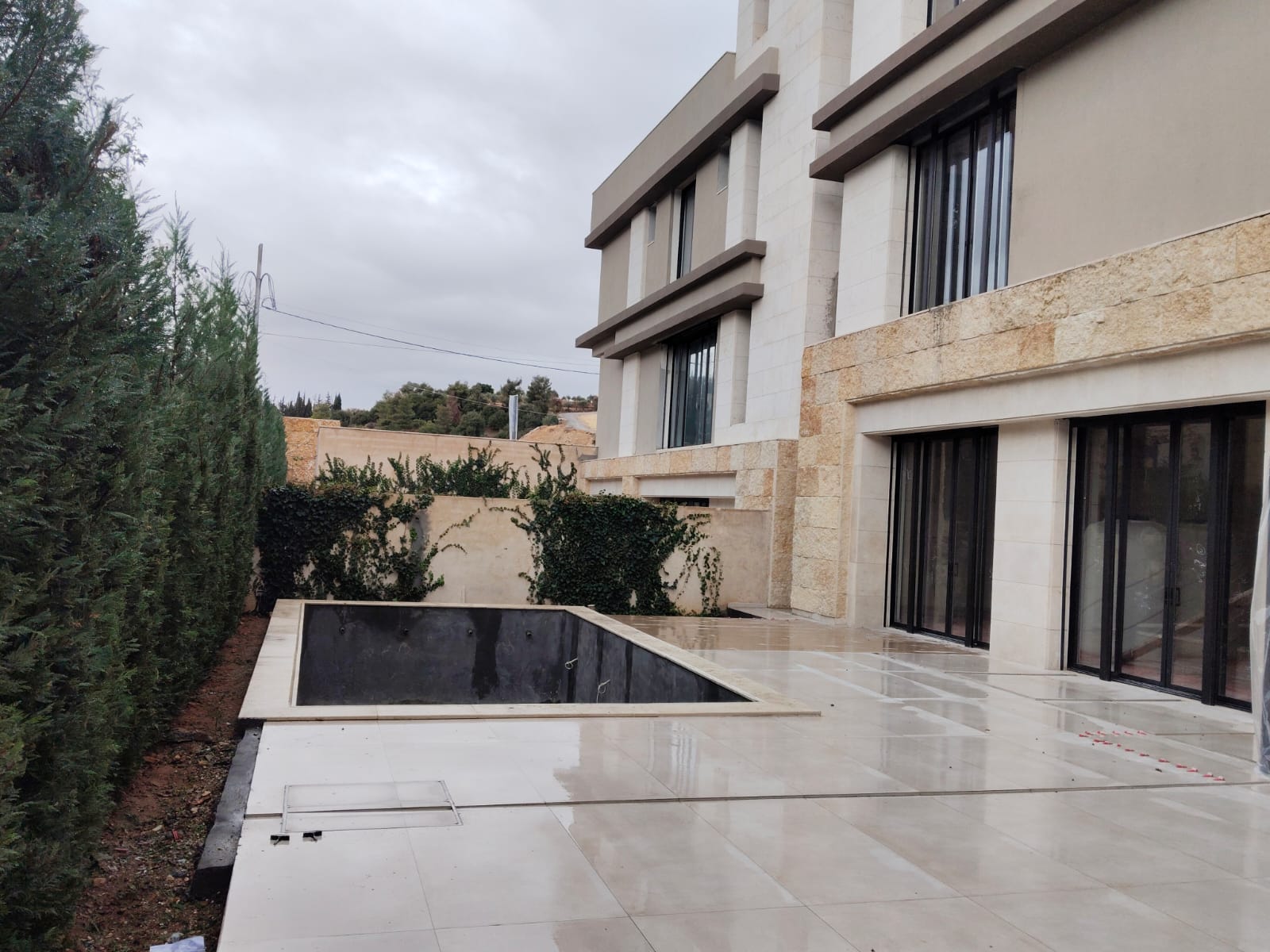 WhatsApp Image 2026-04-08 at 23510 PM 8 Modern beige, multi-story house with large glass doors opening to a tiled patio and an empty rectangular pool, overcast sky above.