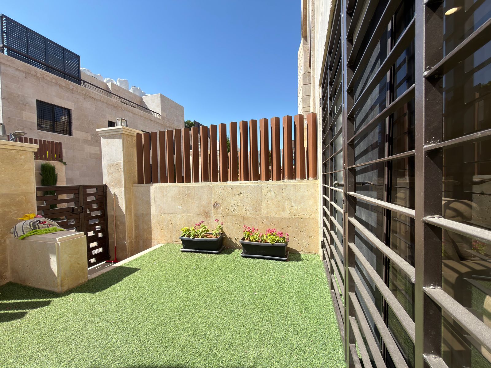a039c182-88b4-4a52-b1b3-f2adbe7d730b Small courtyard with artificial grass, two planter boxes with flowers, beige stone walls, and a brown wooden slat fence behind. Safe outdoor space under a clear blue sky.