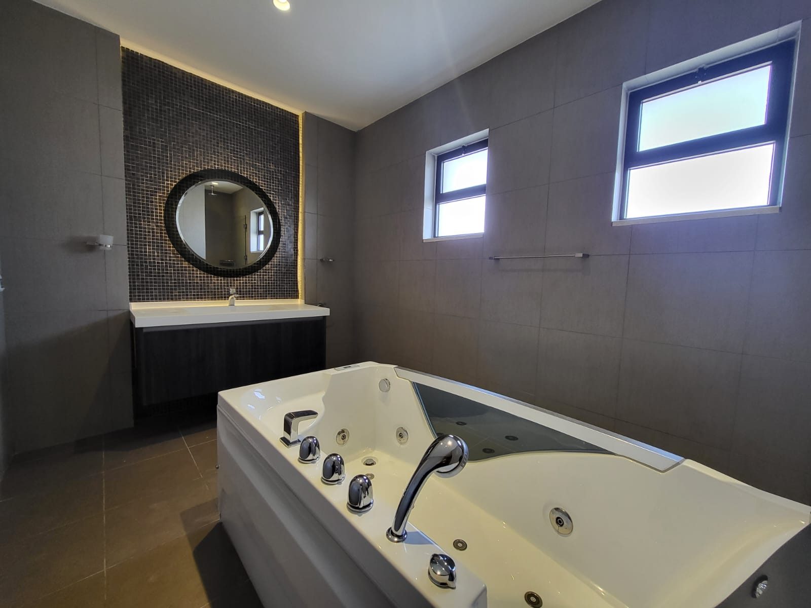 a109fc9b-7704-4d2b-ba2a-d0b93bcb68a5 Modern bathroom with a large white whirlpool tub, dark wood vanity, and round mirror on a mosaic-tiled wall.
