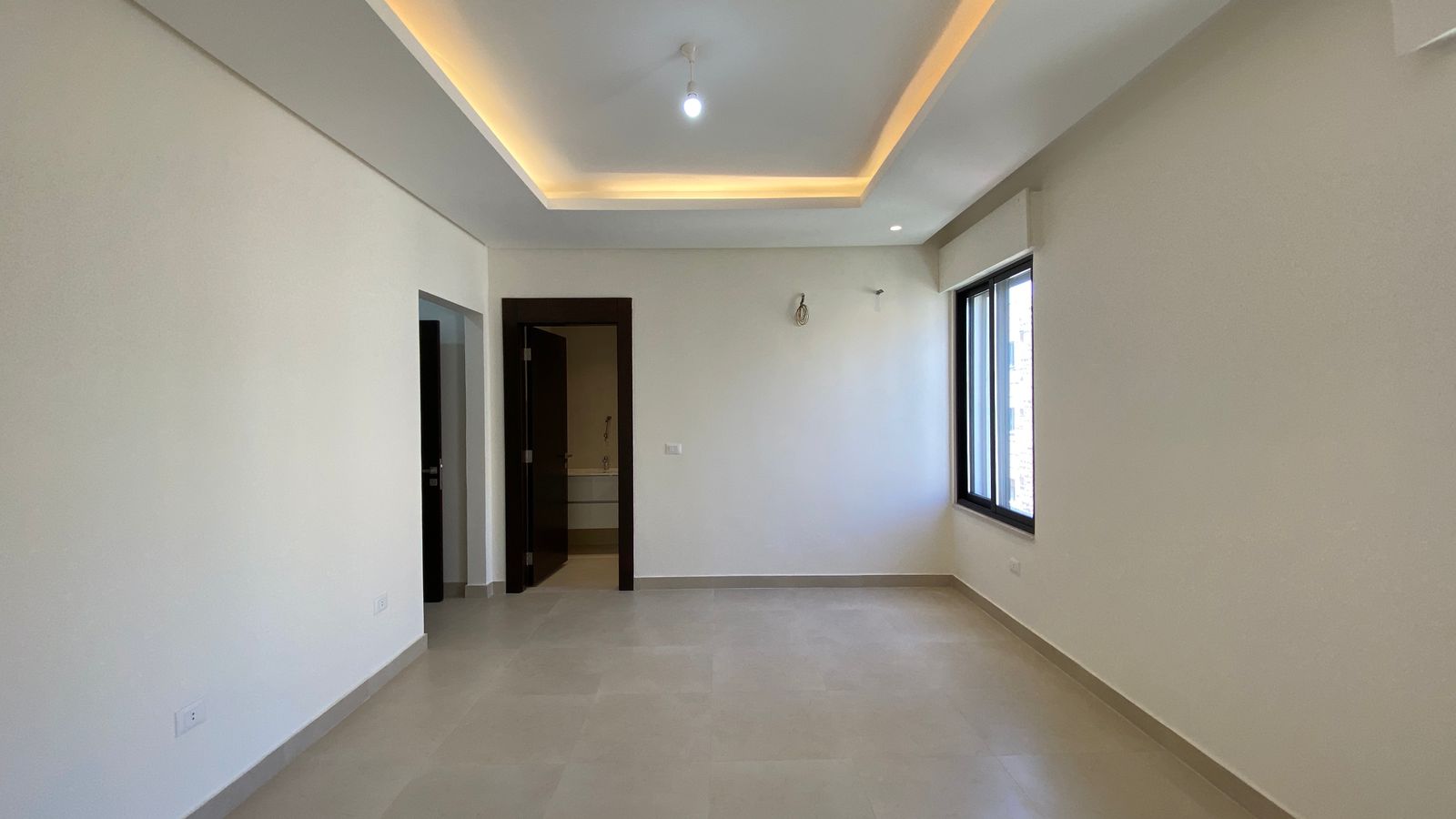 a19ca2cf-f2ca-48f5-9d96-388fd25fd3dd Empty modern living room with a recessed lit ceiling, light tiled floor, and a large window on the right side.