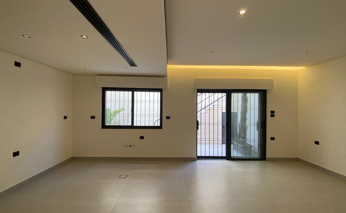 Empty modern living room with tiled floor, white walls, recessed ceiling lights, a barred window, and a sliding glass door to a courtyard.