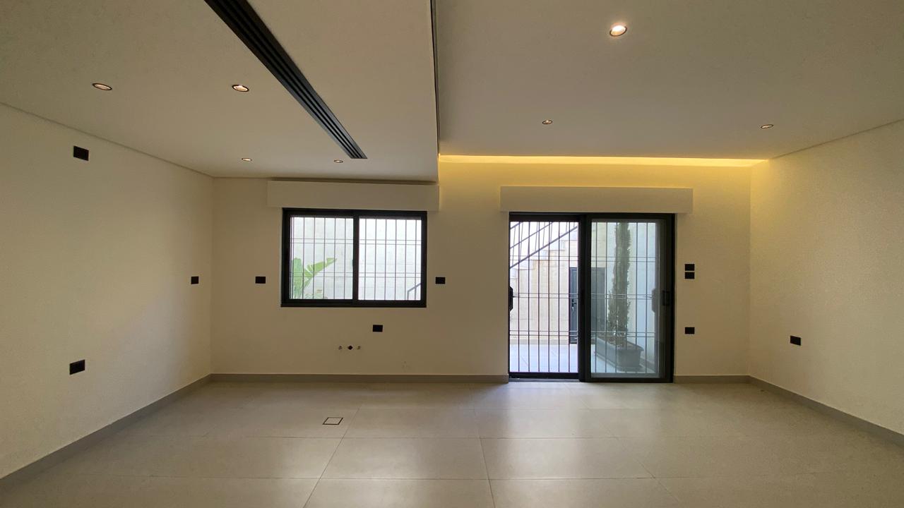 a3a456fd-11e8-40b1-9967-ba7b46a5a2a4 Empty modern living room with tiled floor, white walls, recessed ceiling lights, a barred window, and a sliding glass door to a courtyard.