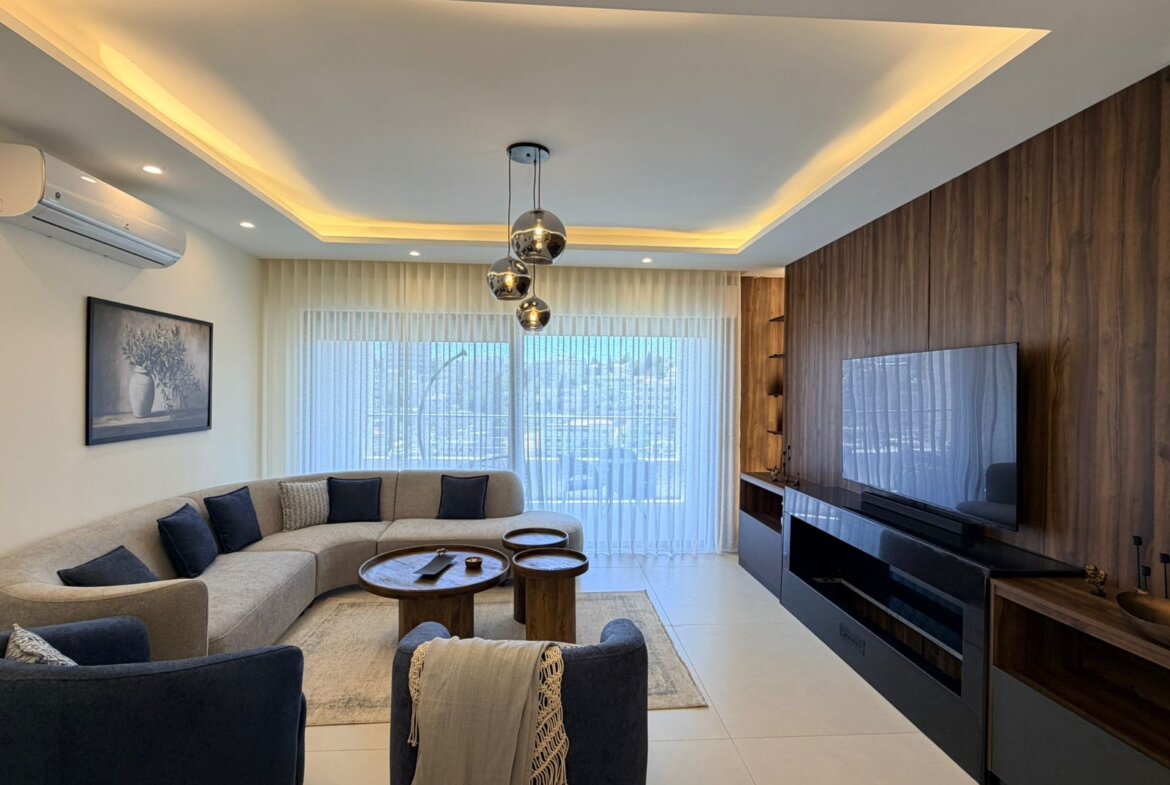 Contemporary living room with a beige L-shaped sofa, navy cushions, and round wooden coffee tables facing a wall-mounted flat-screen TV on a dark wood panel wall.