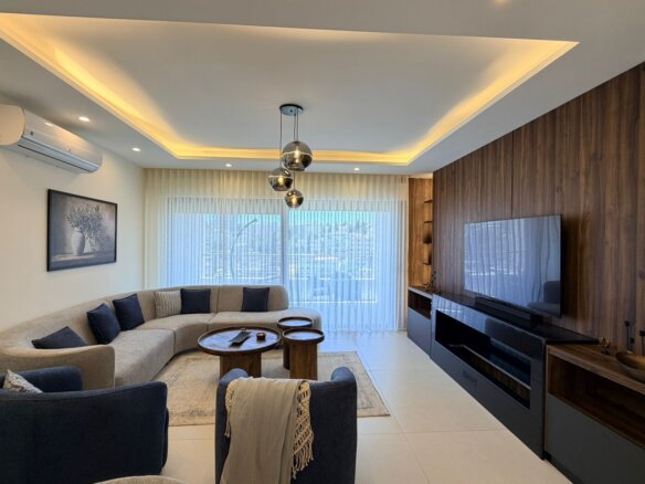 Contemporary living room with a beige L-shaped sofa, navy cushions, and round wooden coffee tables facing a wall-mounted flat-screen TV on a dark wood panel wall.