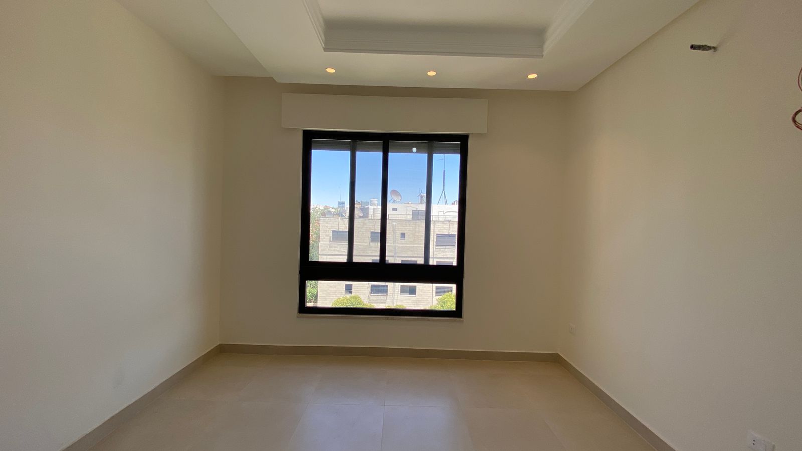a4674016-fefc-4116-9d36-f415a07b51ad Empty beige-walled room with a large black-framed window overlooking a cityscape.
