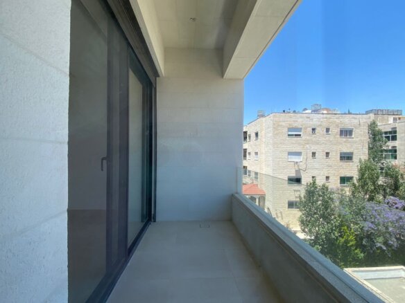 Empty balcony with white tiled floor and glass sliding door, overlooking rooftops and trees under a clear blue sky.
