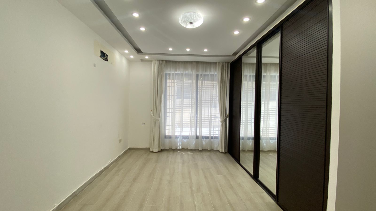 a61aaad5-12da-45a9-a6fe-5482cf1bf061 Empty modern room with large window and sheer white curtains, plus a dark sliding-door wardrobe with mirrored panels on the right.”