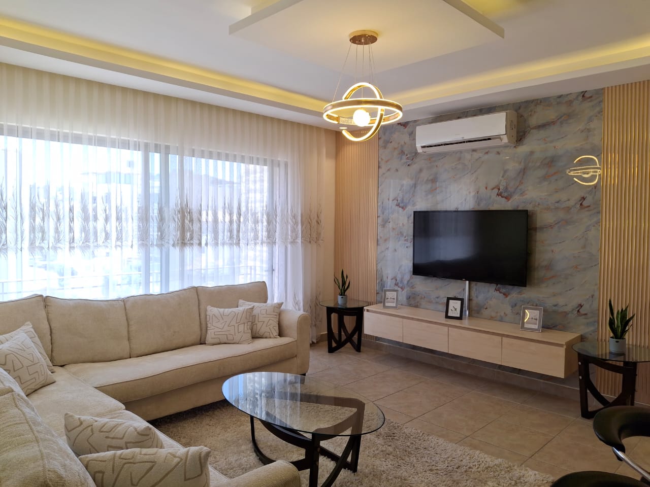 a8e965ef-2789-4785-9d7d-ba615fe5e022 Modern living room with a beige sectional sofa, glass coffee table, and a marble feature wall with a mounted TV.