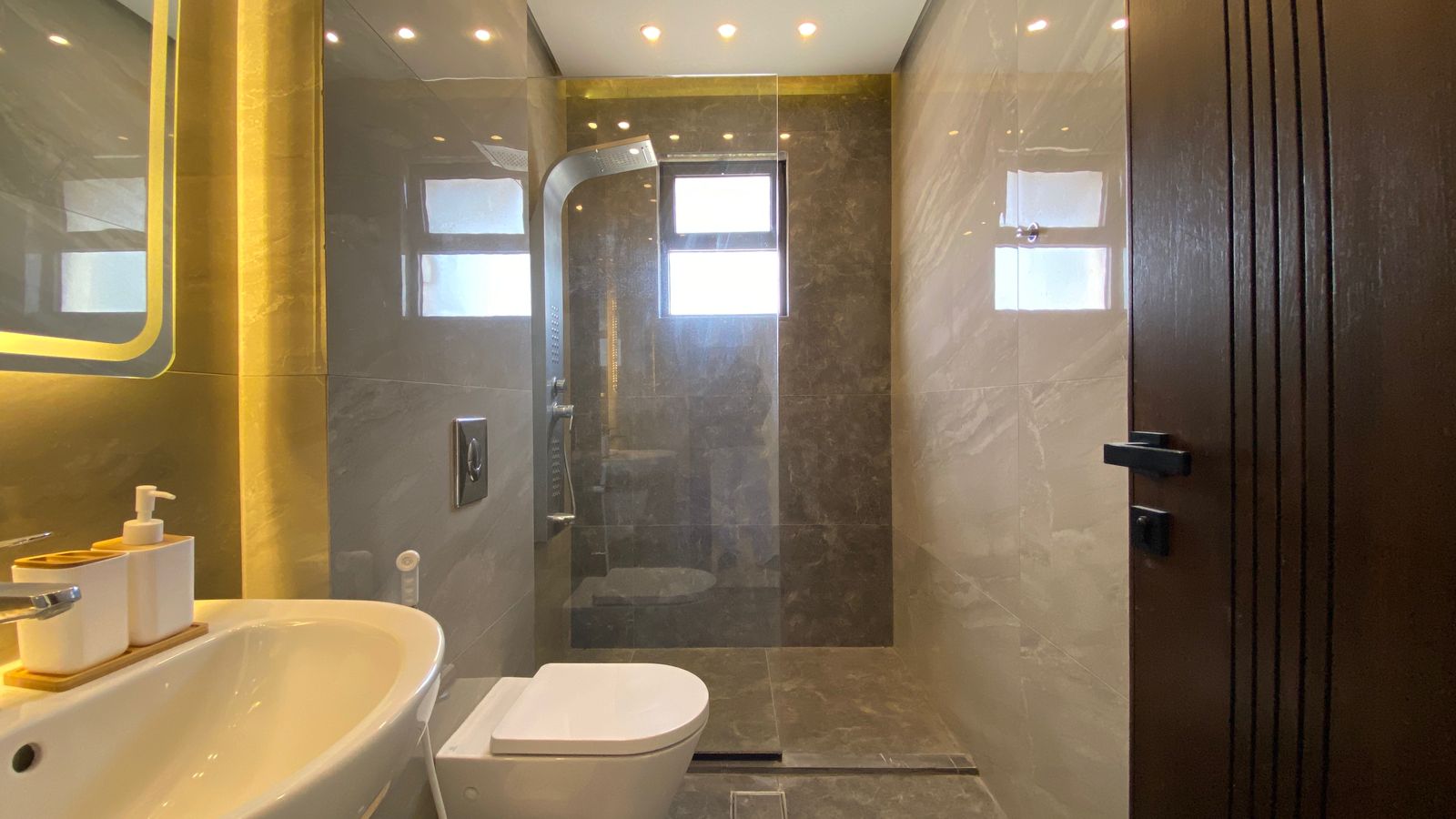 a9a46fa6-caf7-449f-a40b-6de3757fcbb3 Modern bathroom with dark marble walls, a glass-enclosed shower at the back, white toilet and sink, and a wooden door on the right.