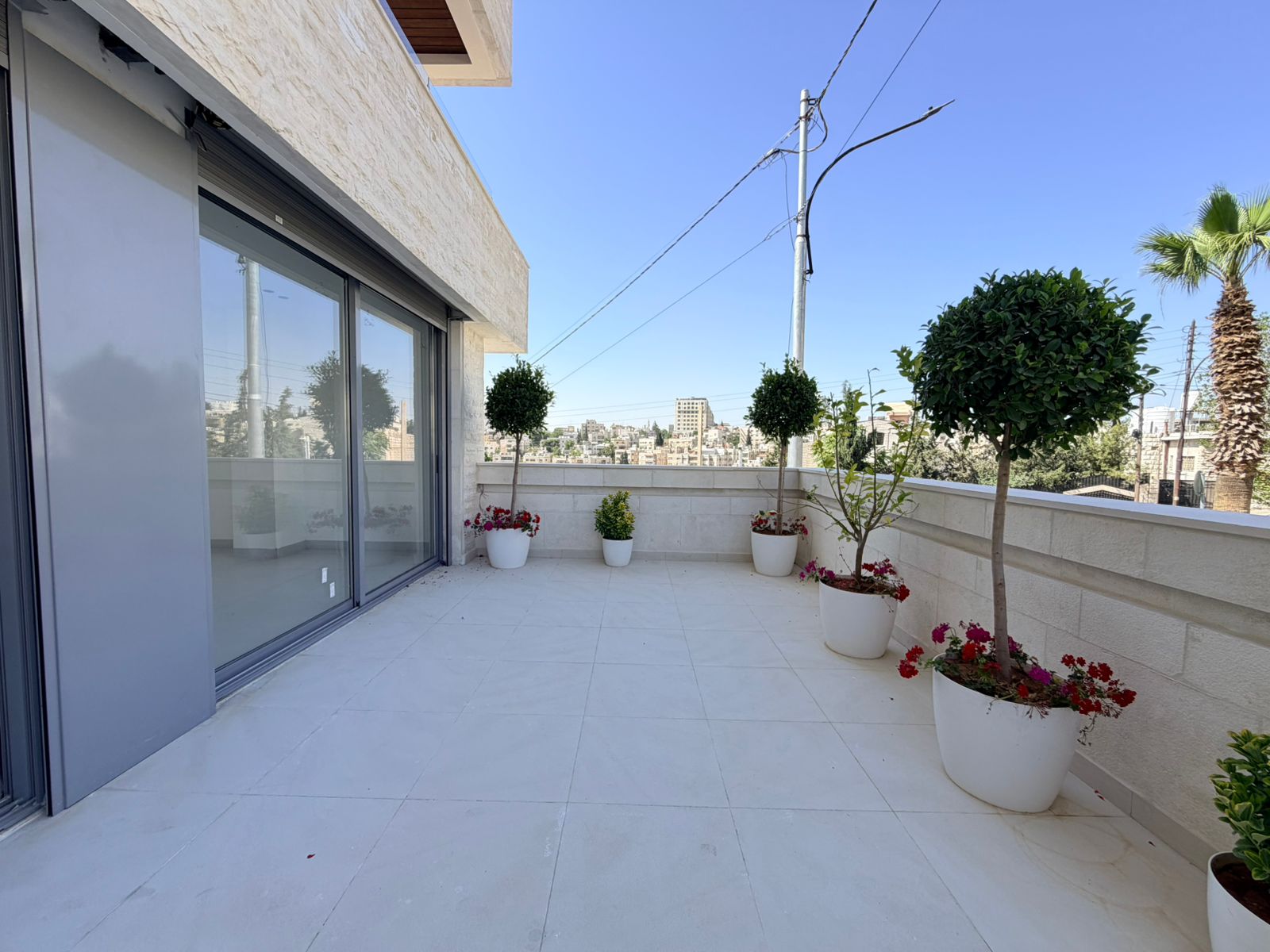 a9d26d9c-e0b9-42bd-98c0-f91c3e9696b2 Bright rooftop terrace with sliding glass doors, white tiled floor, and potted trees against a low wall overlooking the city.