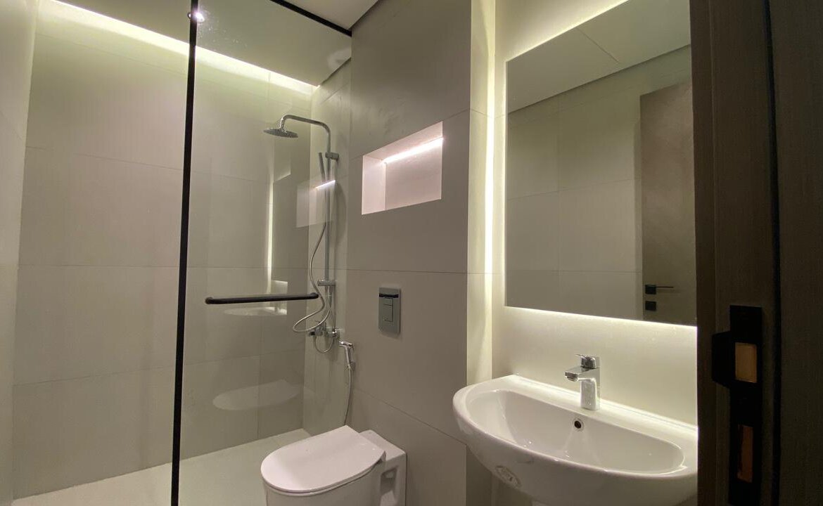 Modern bathroom with a glass-enclosed shower, wall-mounted toilet, and a white sink beneath a backlit rectangular mirror.