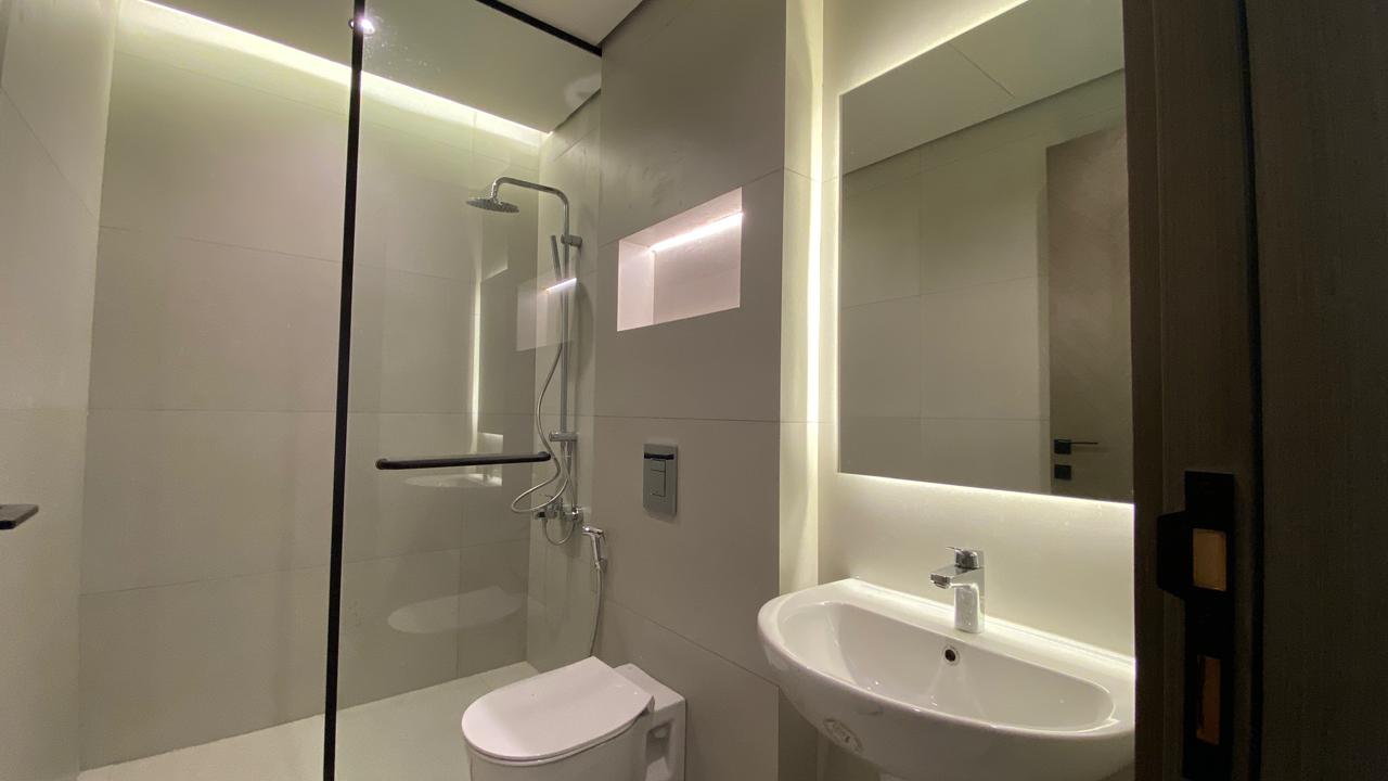 a9d9ccd5-1808-4804-a22a-3aef799894a7 Modern bathroom with a glass-enclosed shower, wall-mounted toilet, and a white sink beneath a backlit rectangular mirror.