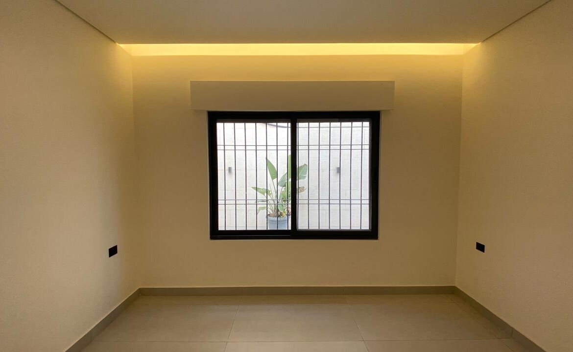 Empty beige room with a barred window; a potted plant is visible outside, under warm ceiling lighting.