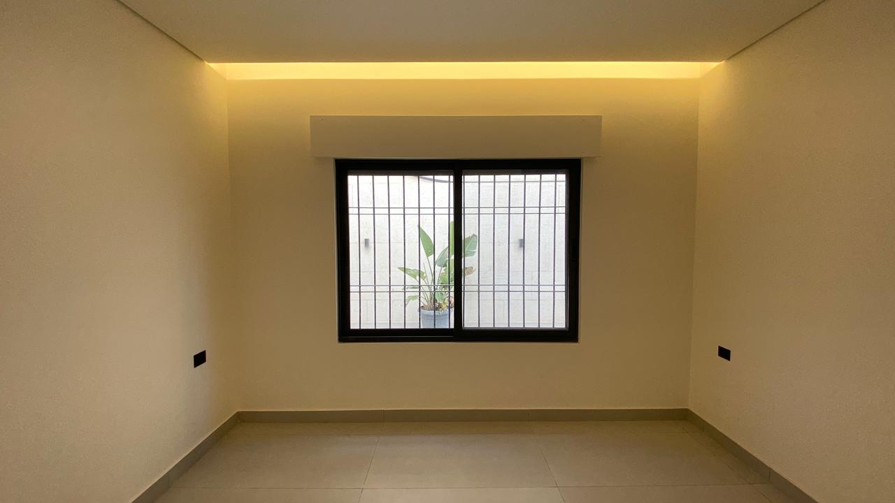 aa931f76-8829-4dd9-a853-cd2d6a5b0c24 Empty beige room with a barred window; a potted plant is visible outside, under warm ceiling lighting.
