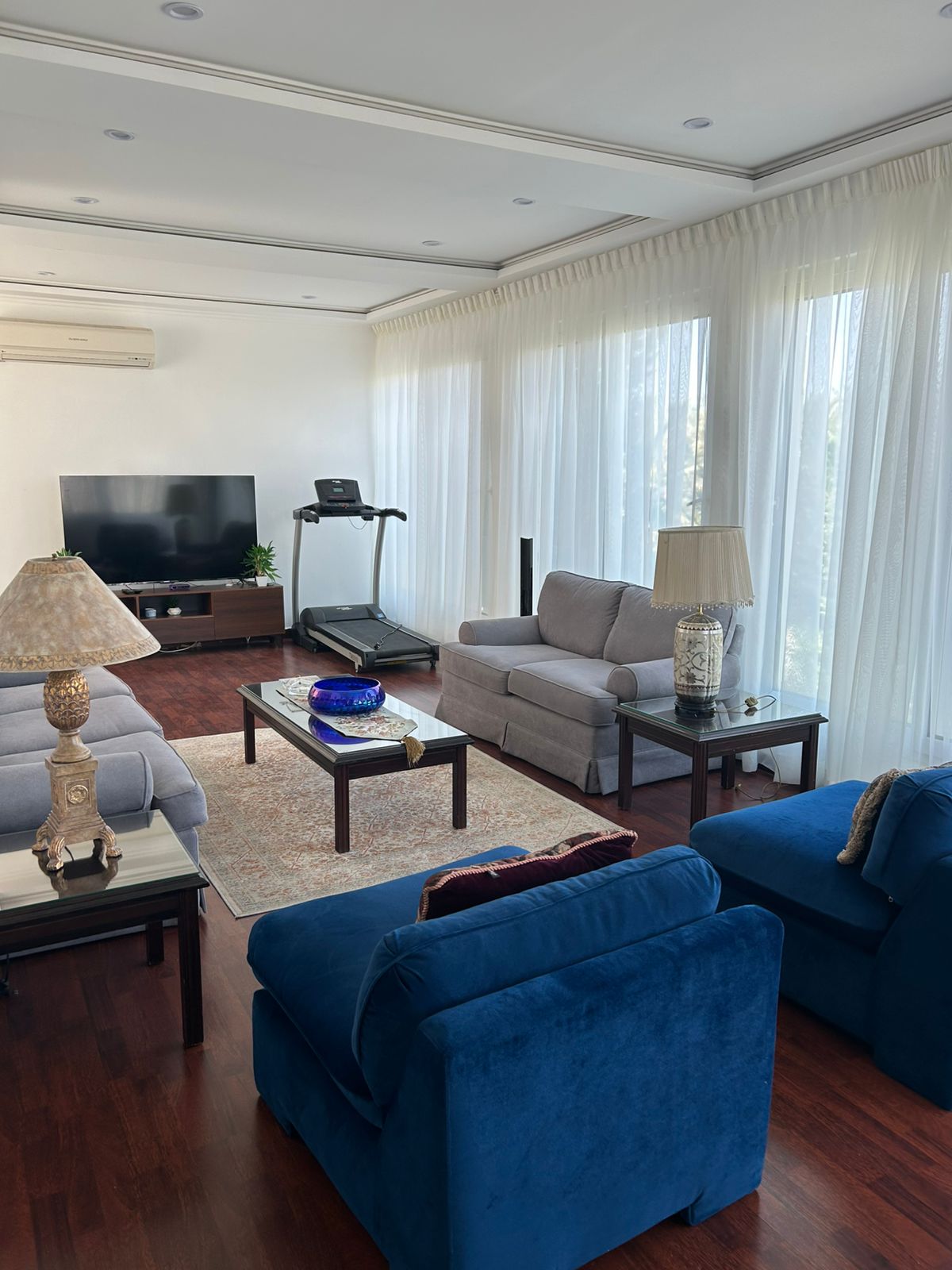 aab275ba-a031-4b26-9086-bcf49f6c7eb5 Bright living room with white sheer curtains, hardwood floors, and a mix of blue and gray sofas around a glass coffee table.