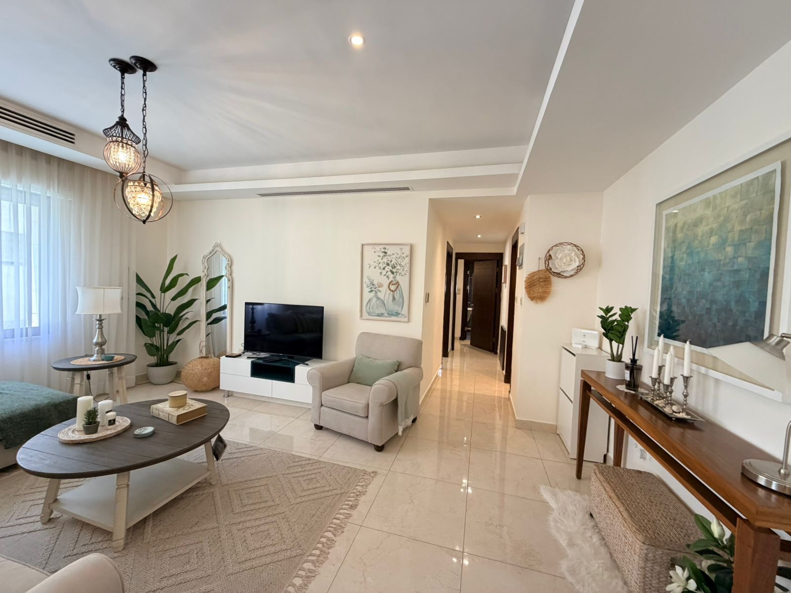 abfa0b30-2eee-42ce-b831-e5e589c82326 Bright living room with beige sofa, round dark wood coffee table, TV on a white console, and large sheer-windowed wall.