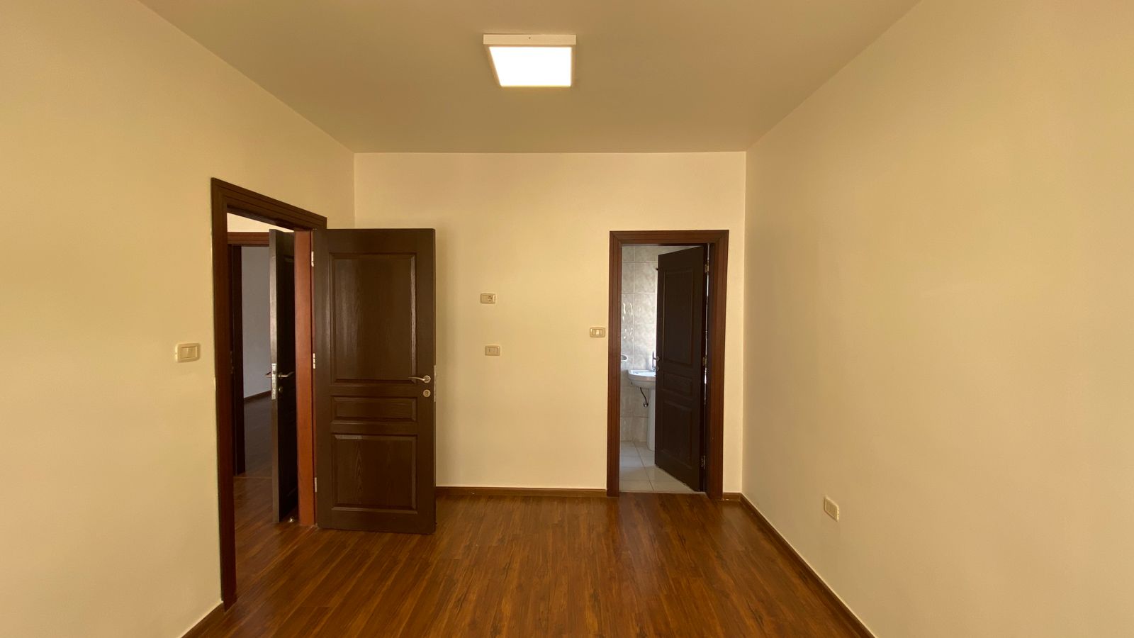 ac0b2f2b-ae91-4898-8e91-5477d1506ce2 Empty rectangular room with warm wood flooring and two dark wooden doors; left door open to another room, right door leads to a tiled bathroom with a sink visible.