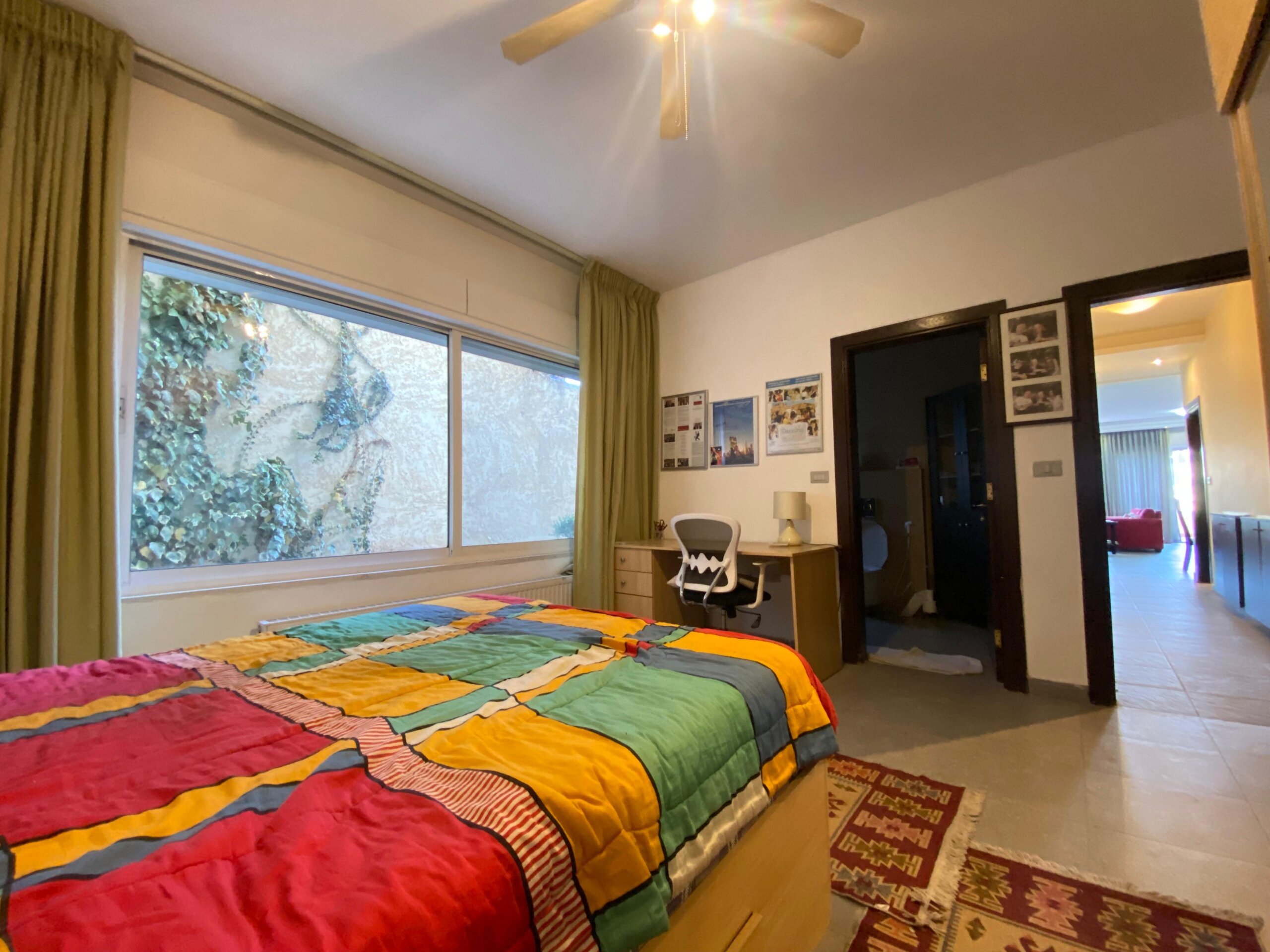 ad488295-da8b-488a-97cb-9e4510dd0dcc Bedroom with two beds covered by rainbow quilts, a desk and chair, and a large window with green curtains nearby.