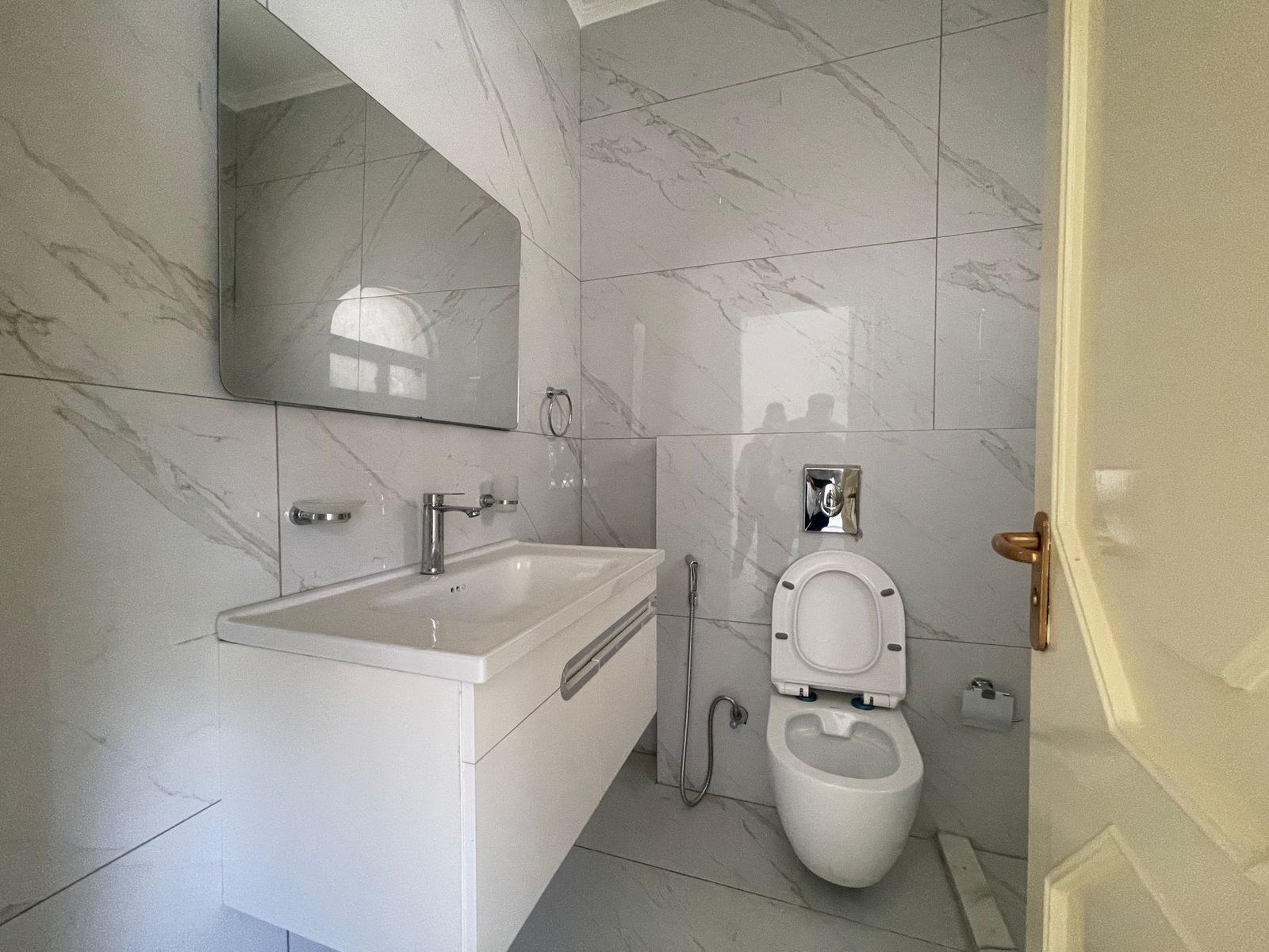 ad67c2cf-ab5d-4c82-a26d-535ee5598894 Bright modern bathroom with white vanity sink, large mirror, marble tiled walls, towel ring, and a wall-mounted toilet near an open door in a sunlit room.