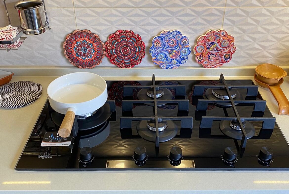 Gas cooktop with four burners; white saucepan with wooden handle on front-left burner, decorative mandala plates along tiled backsplash in the background.