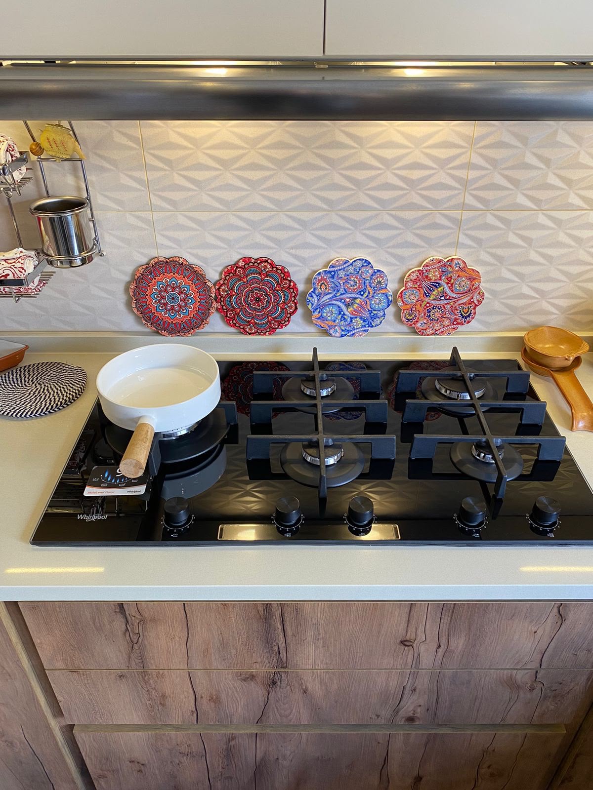ad7553ed-89a9-4c85-9bcf-f22216eea0c7 Gas cooktop with four burners; white saucepan with wooden handle on front-left burner, decorative mandala plates along tiled backsplash in the background.
