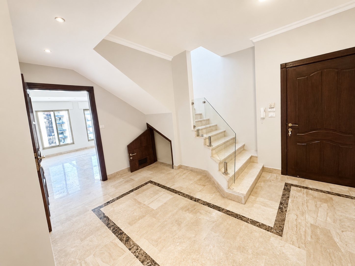 ade36e63-55a7-41c1-9da9-31a3c3810645 Bright foyer with marble floor, stairs with glass railing, and a dark wooden front door on the right; doorway to a sunlit living area on the left.
