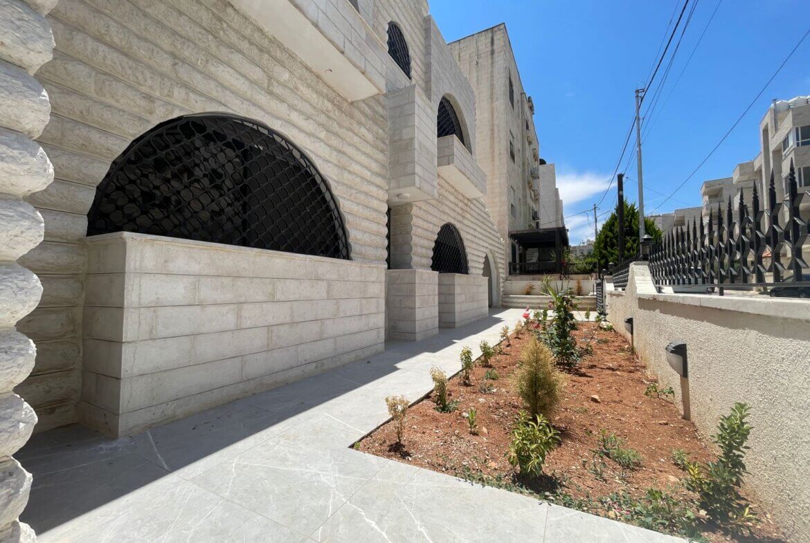 Stone building with arched windows protected by black lattice grilles; a landscaped path runs along a low stone wall.