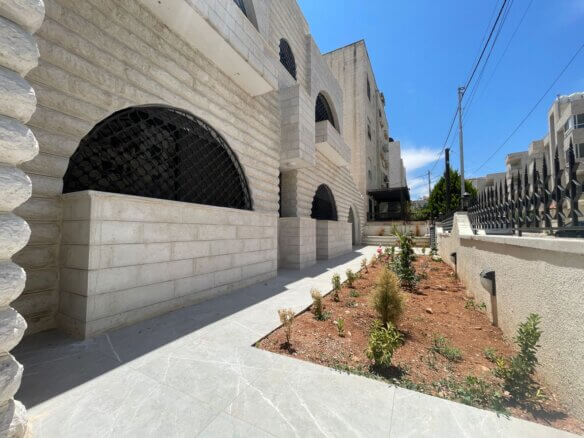 Stone building with arched windows protected by black lattice grilles; a landscaped path runs along a low stone wall.