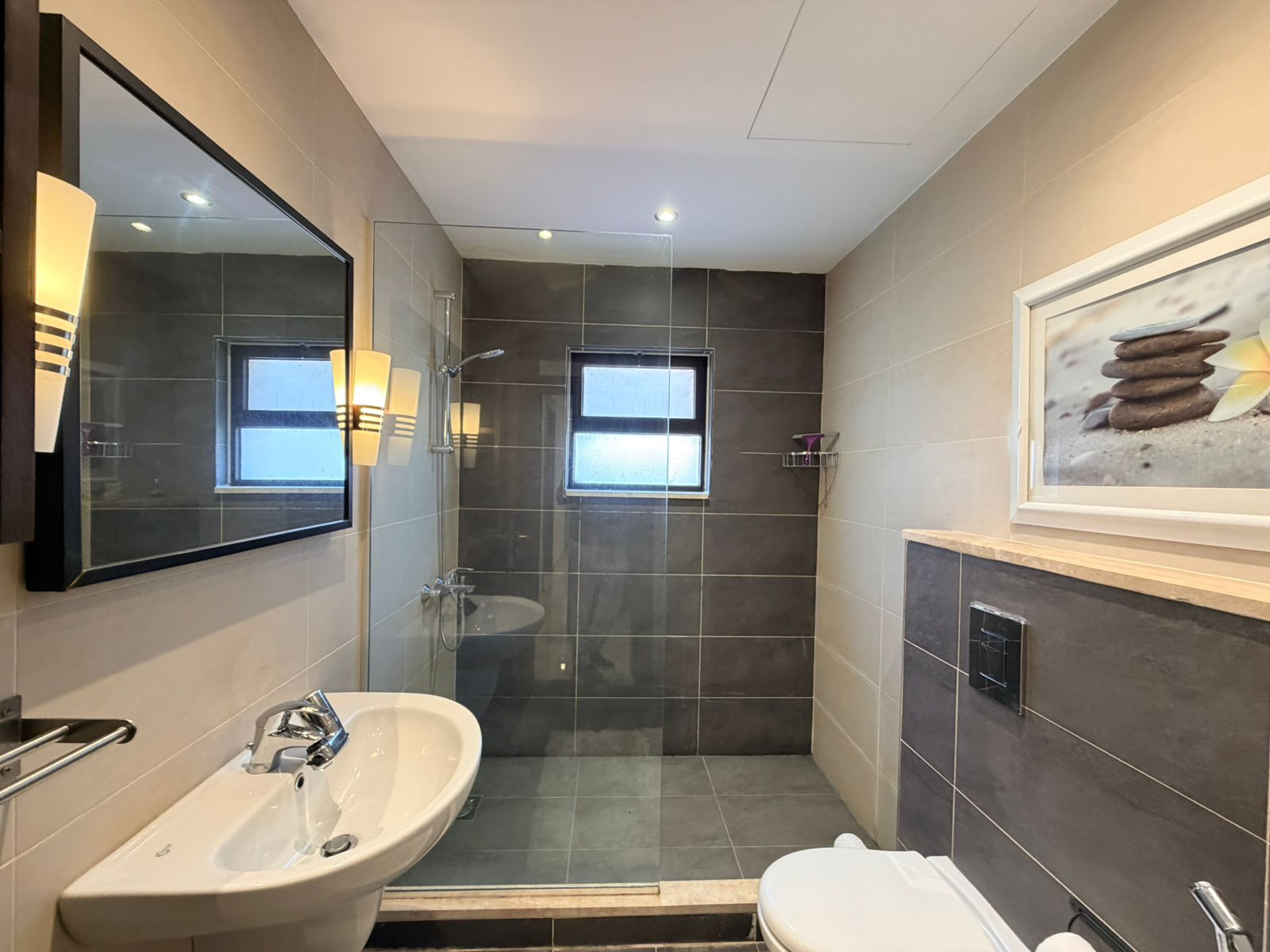 b274203f-9b5e-4f0f-a5c0-1736d8735d49 Modern bathroom with a white pedestal sink, large rectangular mirror, wall sconces, glass shower enclosure, and a framed stone art print on the wall.