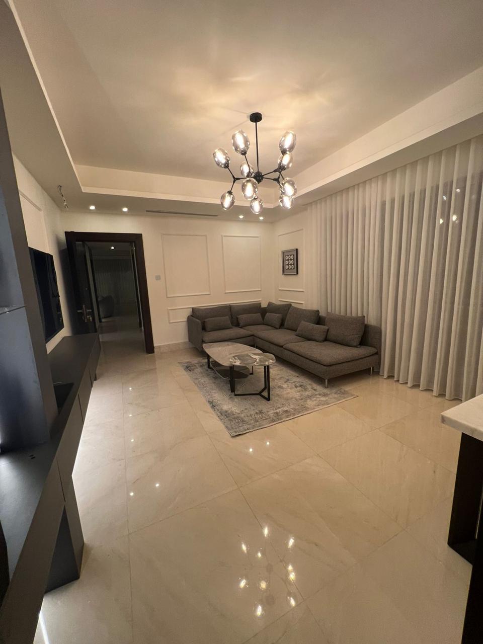 b2ee449f-dff9-4f0a-8860-9ee89ccdbc78 Modern living room with a gray sectional sofa, marble coffee table, and a light area rug under the seating area near white paneled walls and sheer curtains.