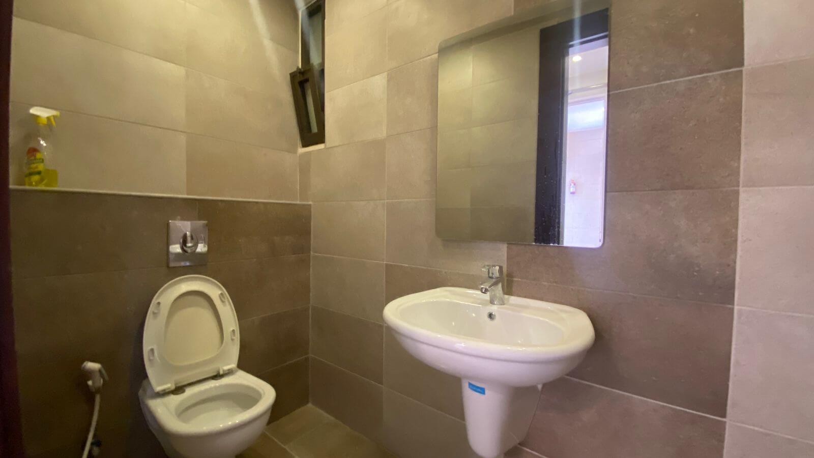 b2ff3316-fb81-46ee-9ab5-f9c51297df57 Bathroom with wall-mounted toilet and pedestal sink, rectangular mirror, and a spray bottle on a shelf nearby.