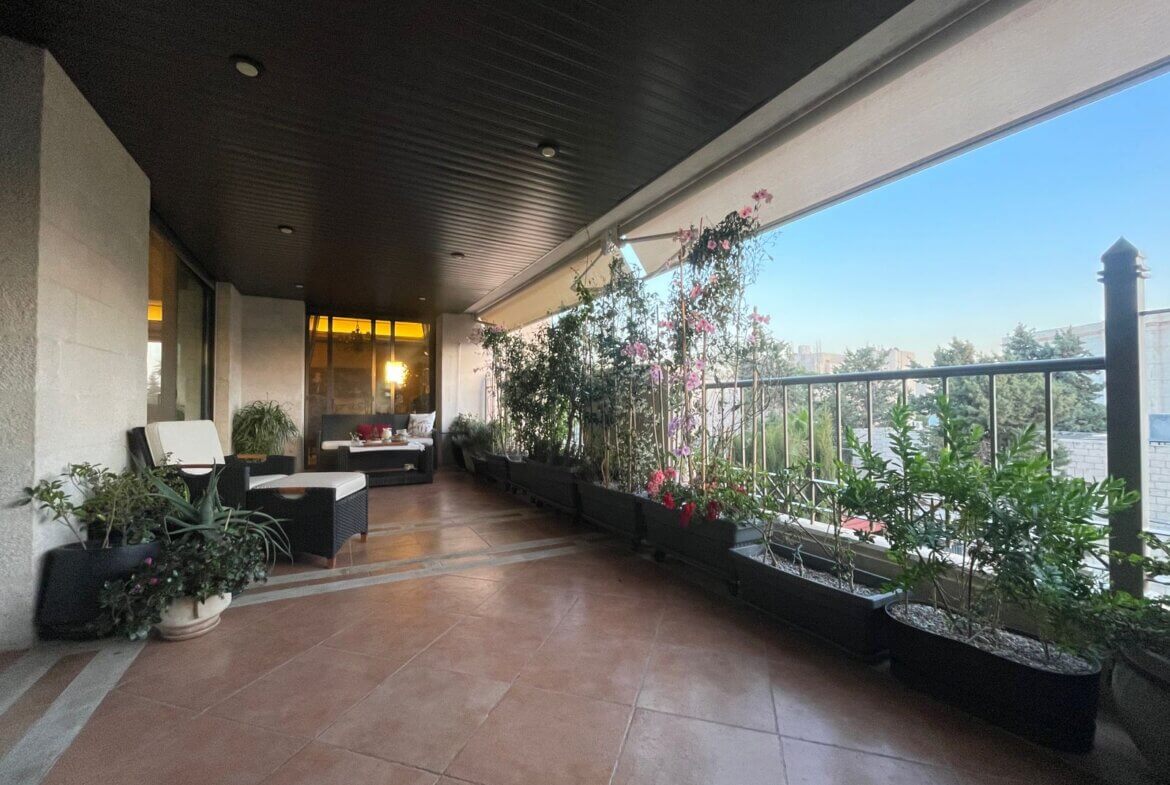 b31c1420-810a-4750-888d-0cc00993d742 Covered balcony with wicker seating, white cushions, and potted plants along a railing, city skyline in the distance at dusk.