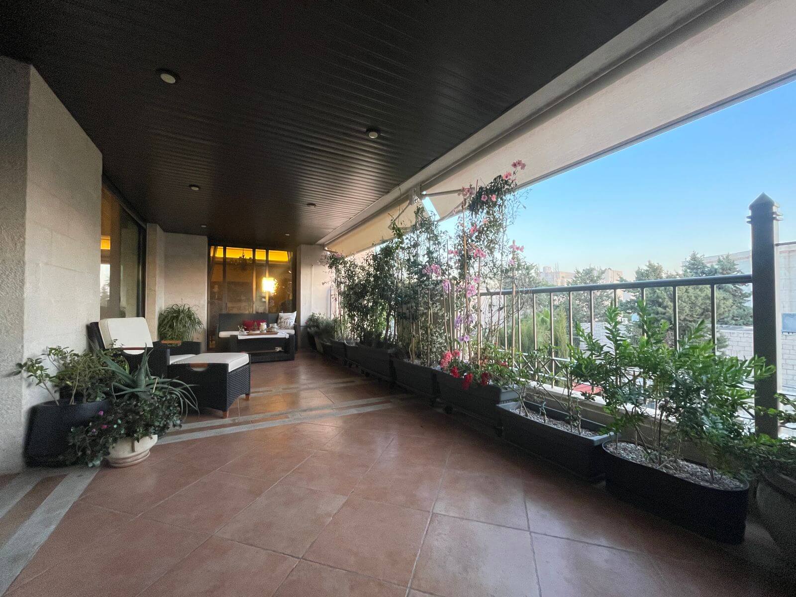 b31c1420-810a-4750-888d-0cc00993d742 Covered balcony with wicker seating, white cushions, and potted plants along a railing, city skyline in the distance at dusk.