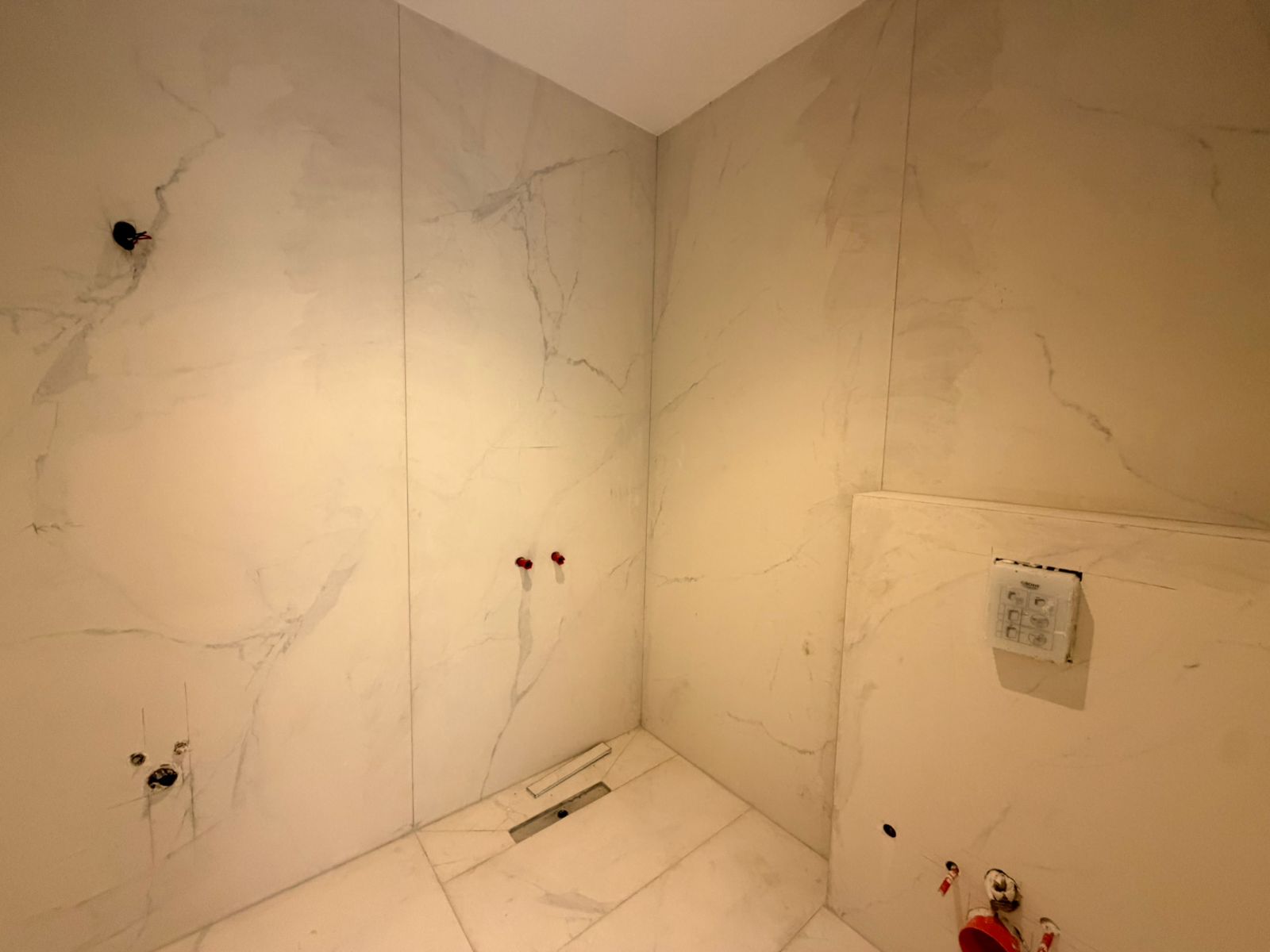 b357df5a-67b8-4f78-a236-ac1ff1a48bcc Unfinished bathroom with large white marble tiles, exposed red plumbing fittings, and a wall-mounted control panel visible on the right.