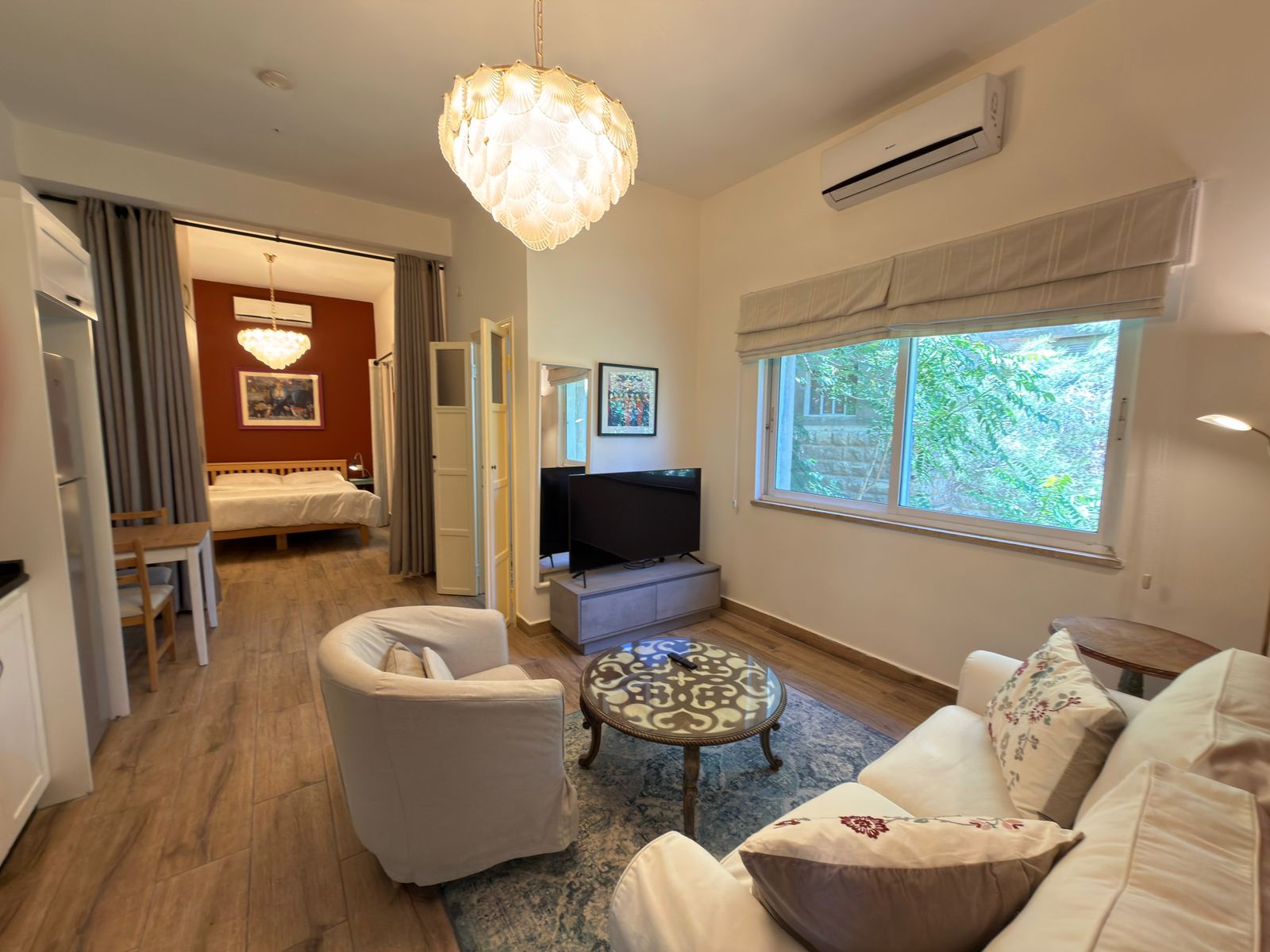 b5212ccd-7eec-49a0-be9d-95389eb3ba3a Living room with white sofa and a patterned round coffee table, facing a TV on a low console and a large window with blinds.