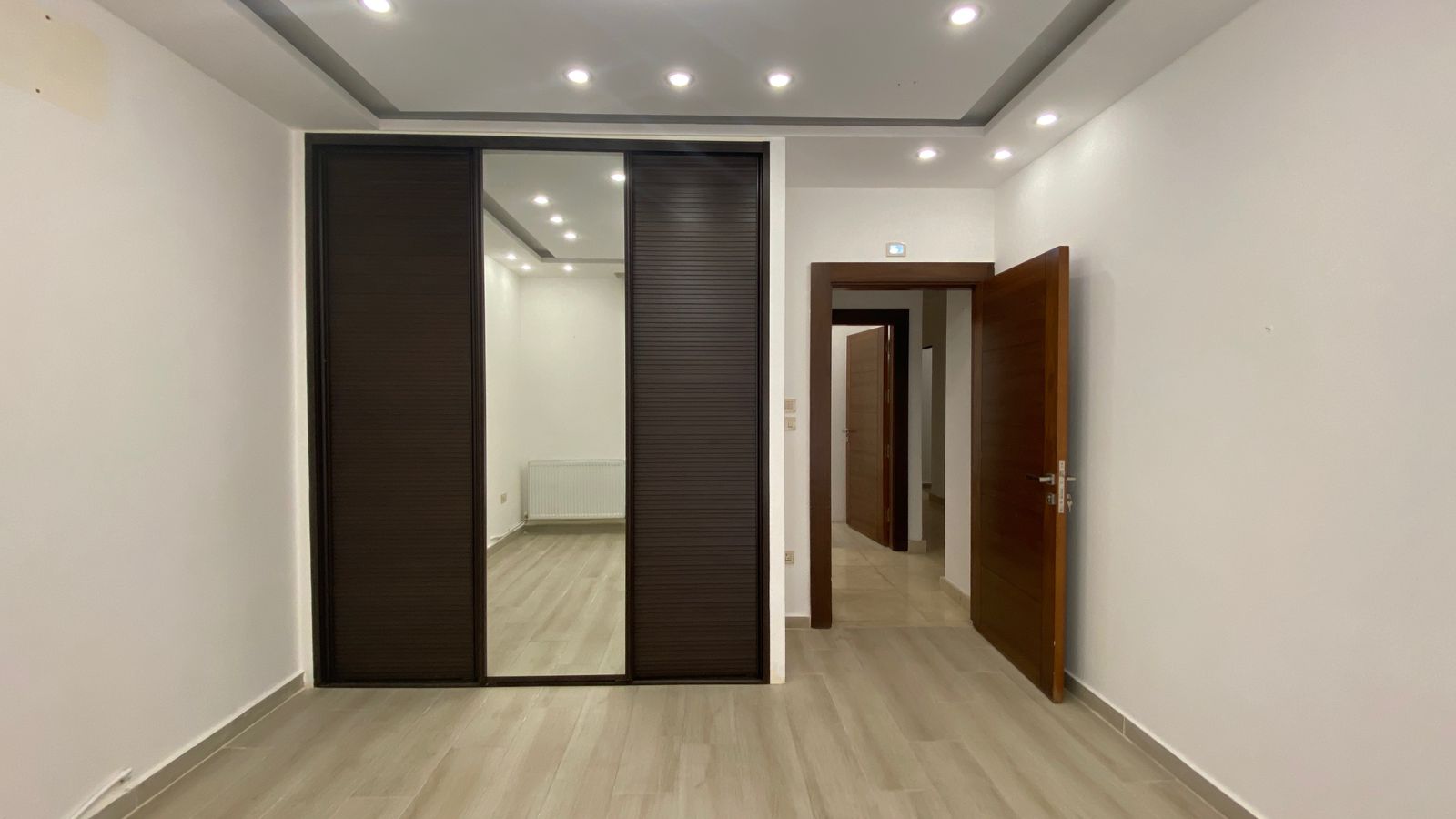 b545b38e-108b-4439-9112-b4349e91d77e Empty modern room with dark wood sliding wardrobe doors, mirrored panel, and an open wooden door to a hallway behind it.