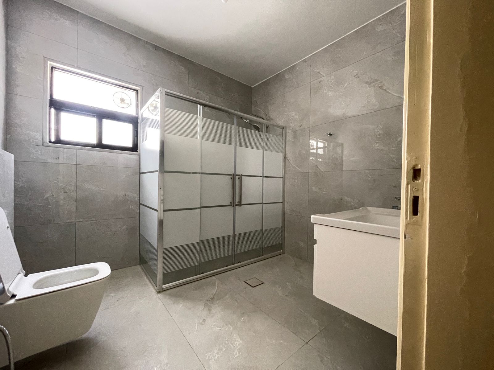 b54b0afc-b6f6-4a2c-83aa-9f84e4f6fce8 Grey tiled bathroom with a frosted glass shower enclosure, toilet on the left, and a white vanity on the right.