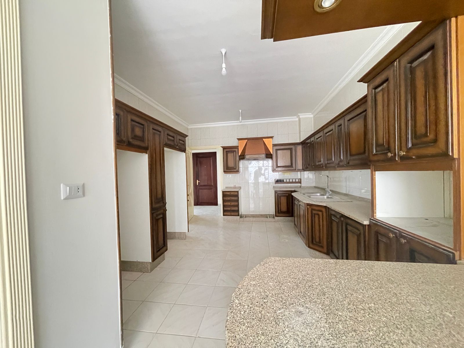 b698fc3e-e5b2-455f-ae17-73b3c352ae96 Spacious kitchen with dark wood cabinets, white tiled walls, and a curved granite island in the foreground.