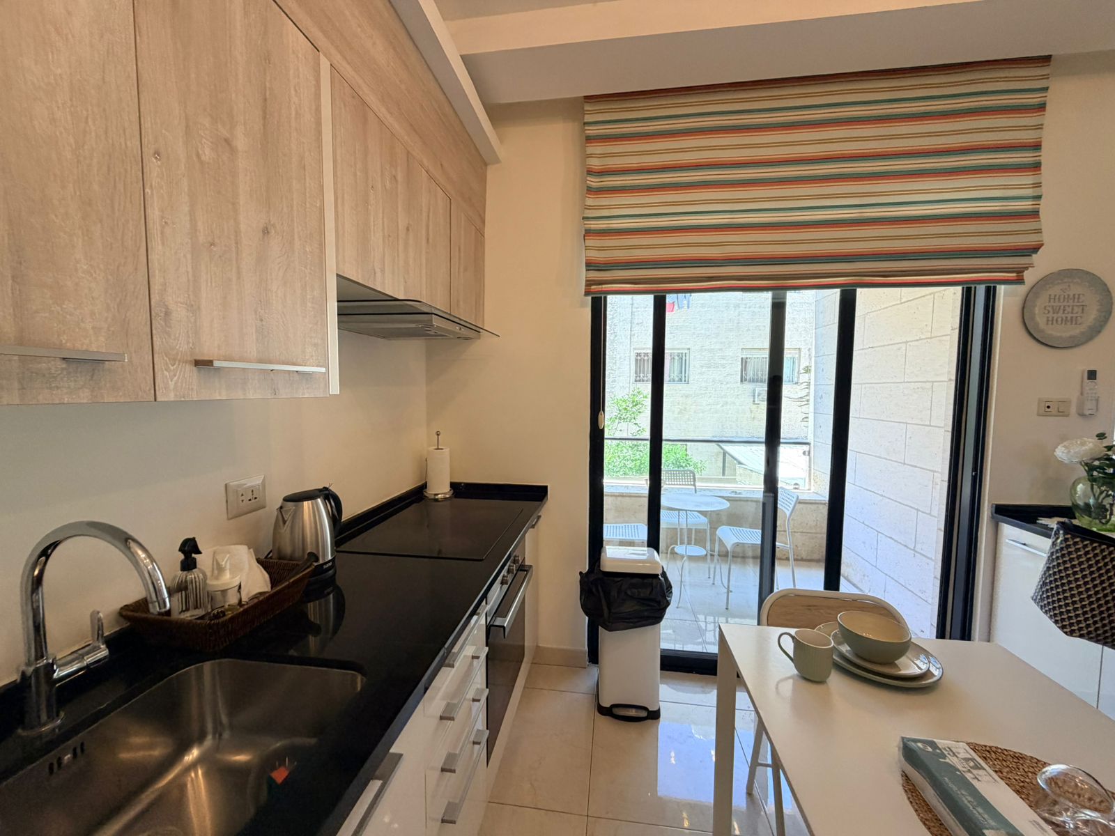 b70dd6c4-9075-4dd3-a4c2-2210ef1fffd4 Kitchen with light wood cabinets, black countertop, and stainless sink; striped window shade and balcony doors in background.