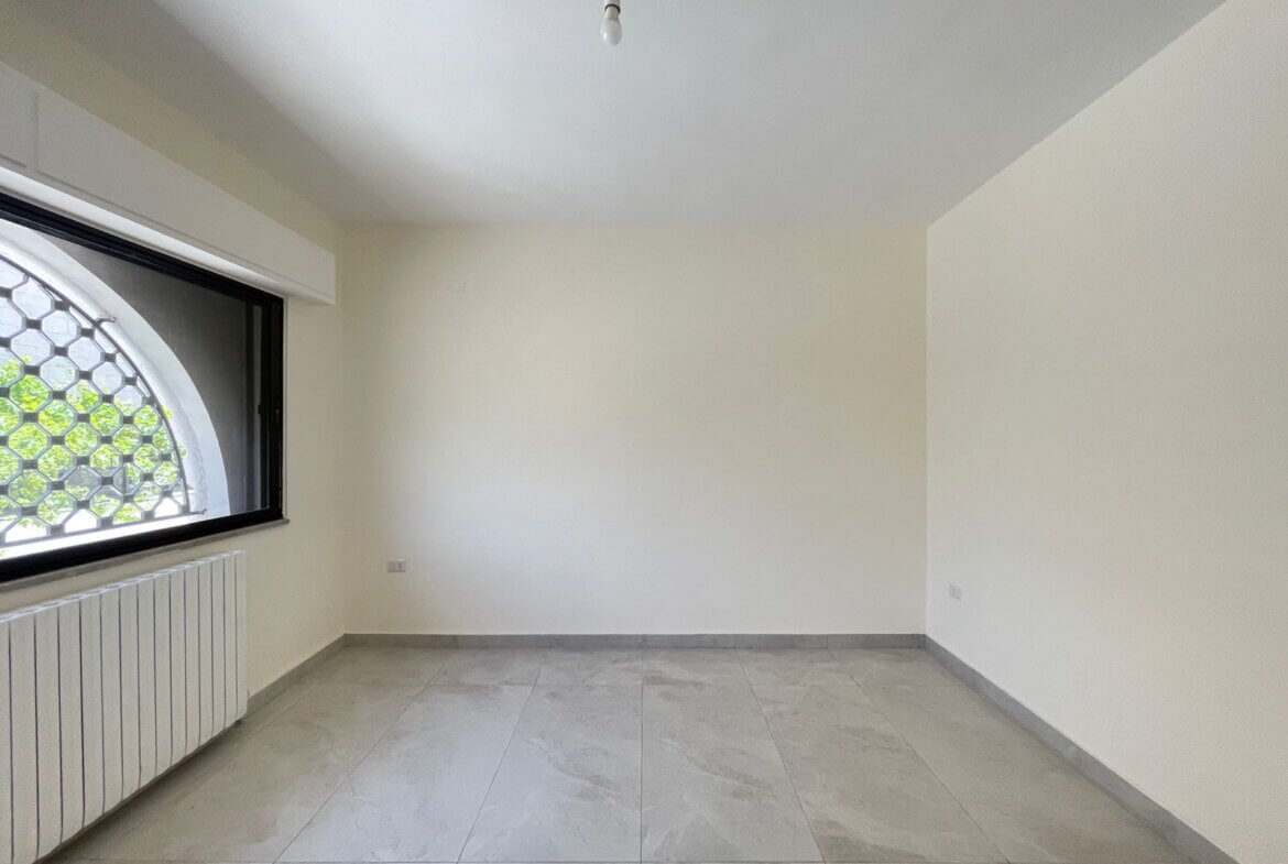 b72f9e52-6771-43f5-a878-97ff153a5ee7 Empty white room with a large arched window and metal grille, a radiator along the wall, and light-gray tiled floor.