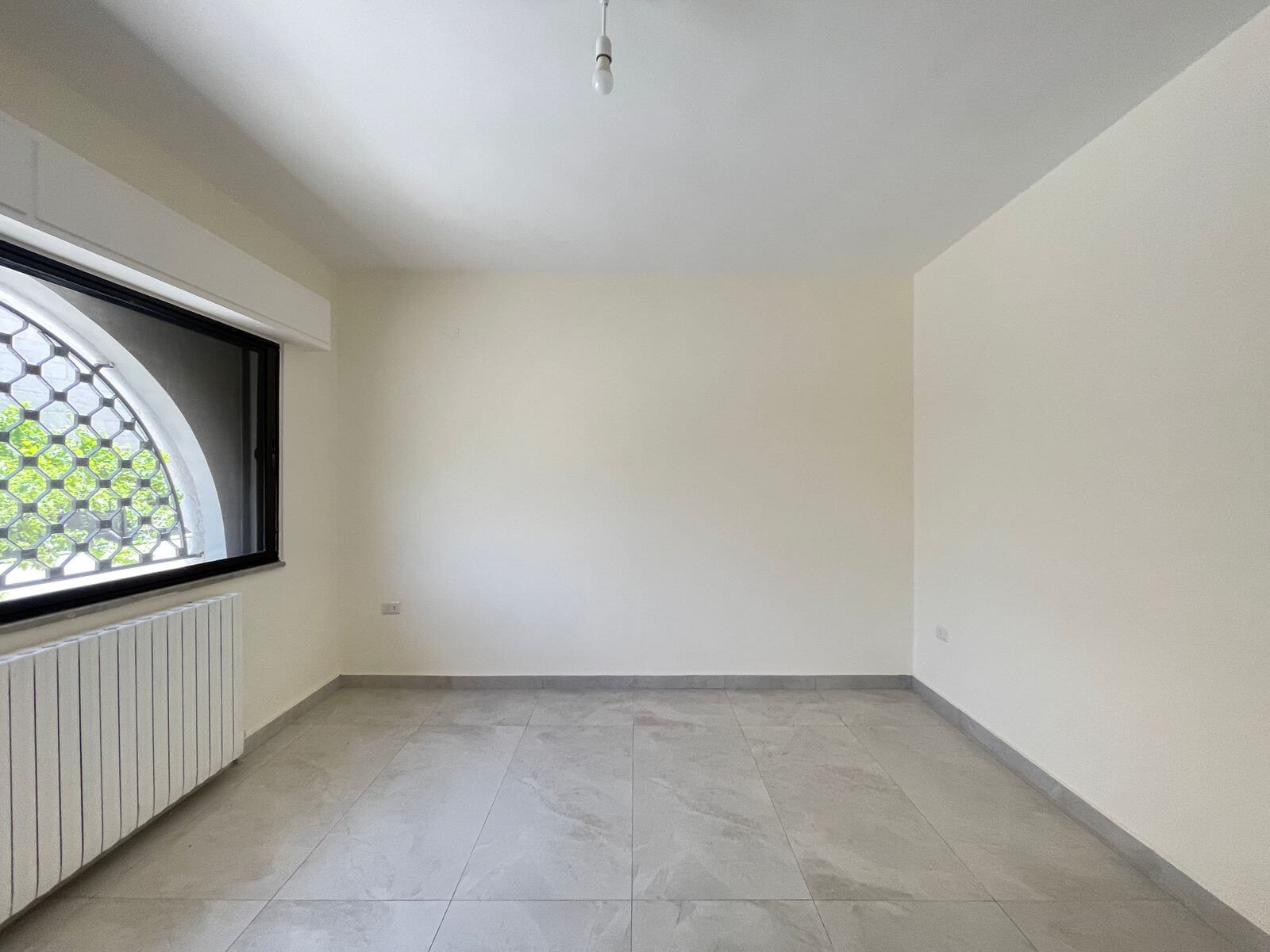 b72f9e52-6771-43f5-a878-97ff153a5ee7 Empty white room with a large arched window and metal grille, a radiator along the wall, and light-gray tiled floor.